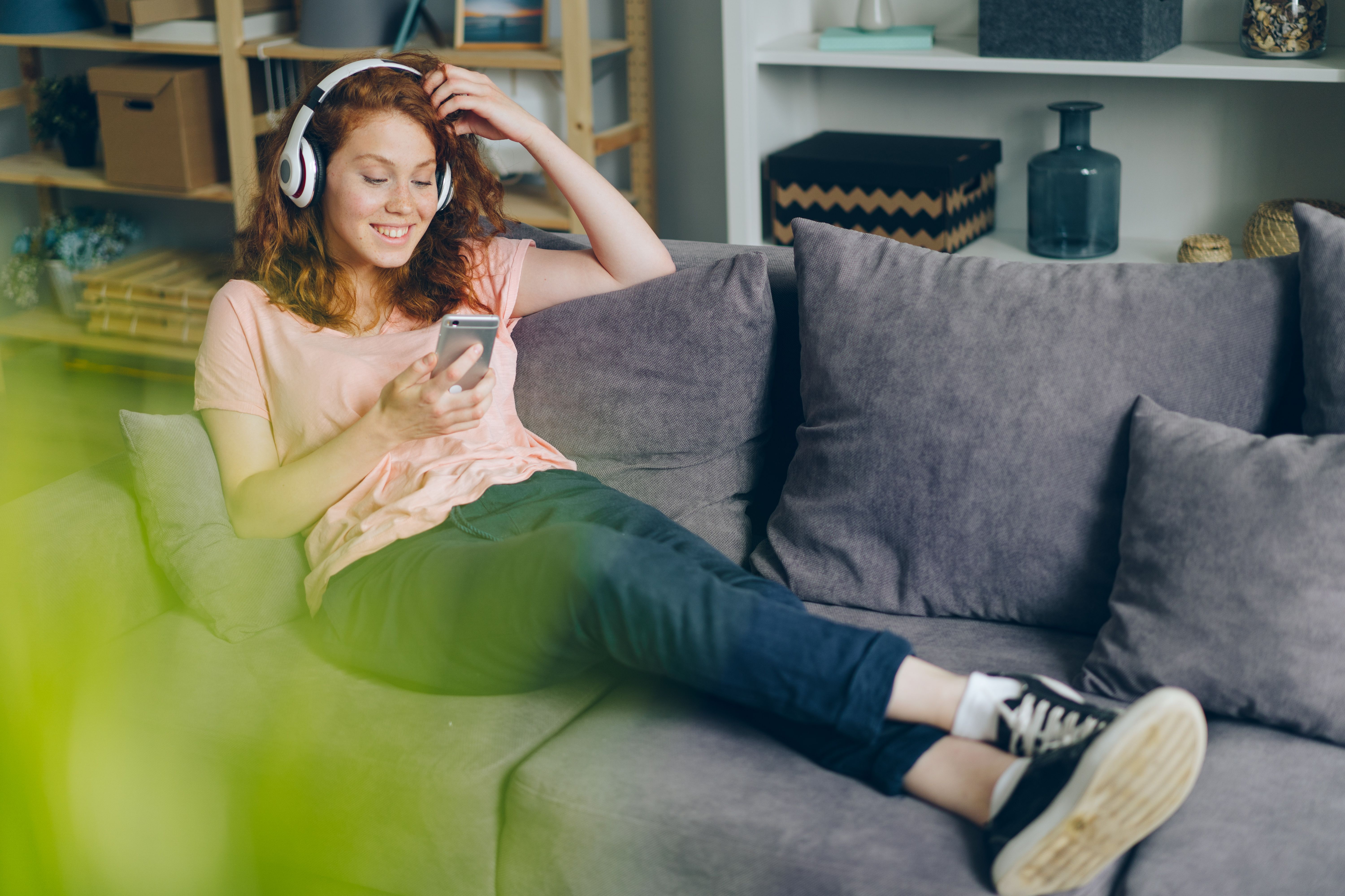 Eine Frau sitzt mit Kopfhörern auf einem Sofa und hält ein Smartphone in der Hand.